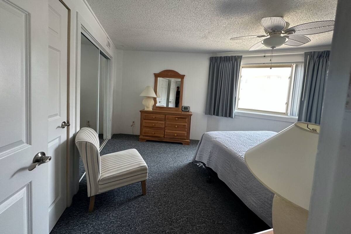 A bedroom with a bed, a dresser, a chair, a ceiling fan, and a window with curtains.