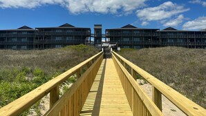 Een houten vogelbeschermingspad dat leidt naar een complex aan het strand met meerdere balkons en een heldere lucht.