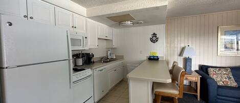A compact kitchen with white cabinets, a microwave, a coffee maker, and a small dining area with a blue couch and wooden chairs.