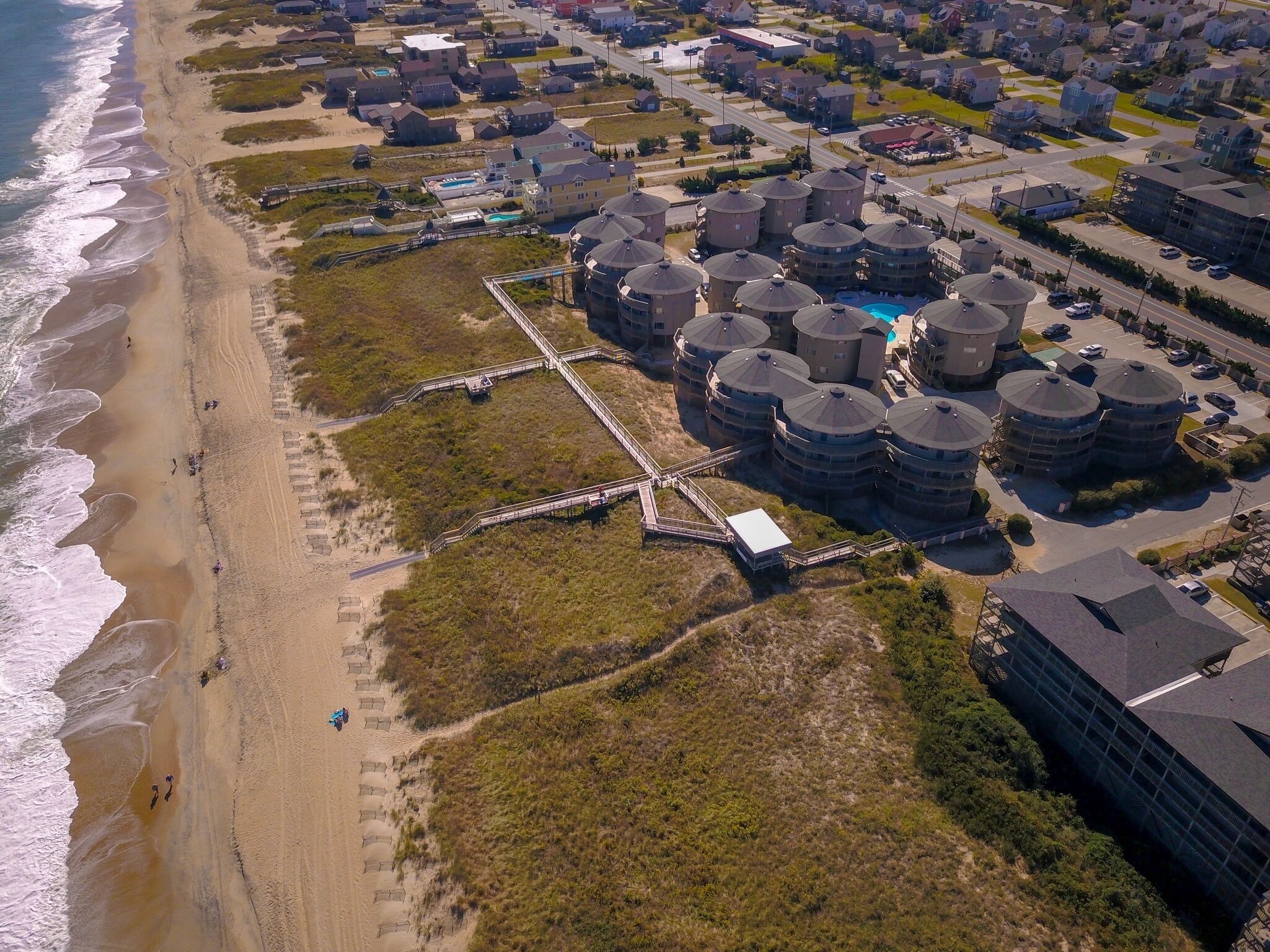 An aerial view of a coastal residential area with multiple cylindrical buildings, a beach, and a pedestrian bridge.