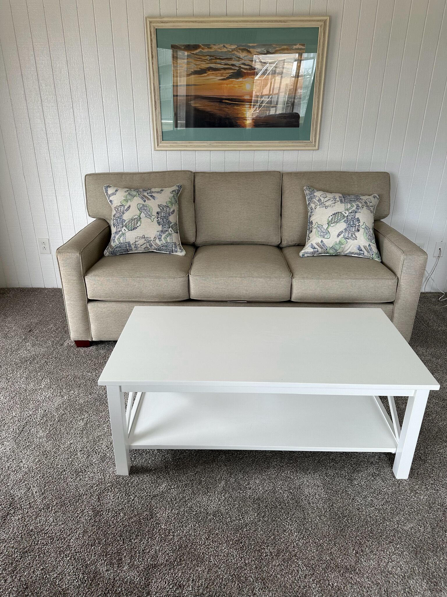 A beige sofa with patterned pillows, a white coffee table, and a framed picture above the sofa.
