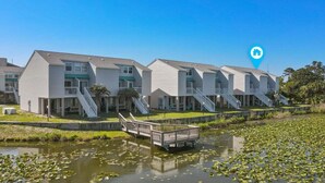 A row of two-story buildings with balconies, a small bridge over a pond with lily pads, and a clear blue sky.