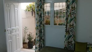 Une pièce avec une porte donnant sur un balcon, une fenêtre avec des rideaux et une petite table sur laquelle est posée une plante en pot.