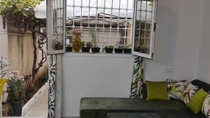 Une pièce chaleureuse avec un canapé vert, un tapis à motifs et une fenêtre donnant sur des plantes et une clôture.