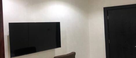 A small room with a flat-screen TV mounted on the wall, a desk with a chair, and a black door.