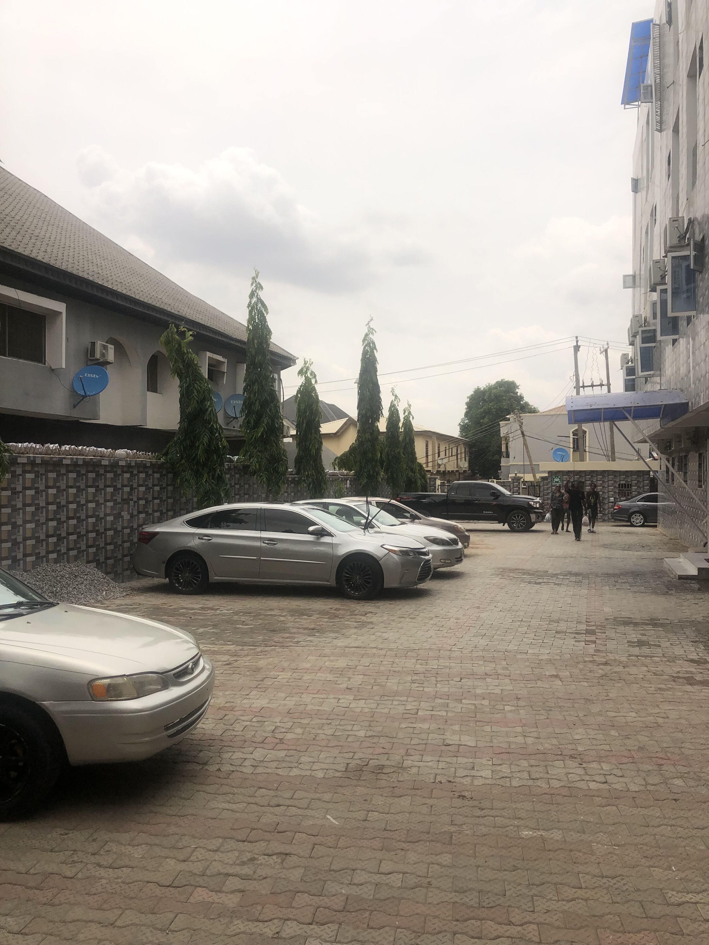 Un estacionamiento con varios autos, un edificio con antenas parabólicas y algunos árboles.