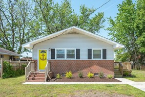 A single-story house with a brick foundation and white siding, a front door with a yellow doorbell, and a small front yard with young trees and shrubs.
