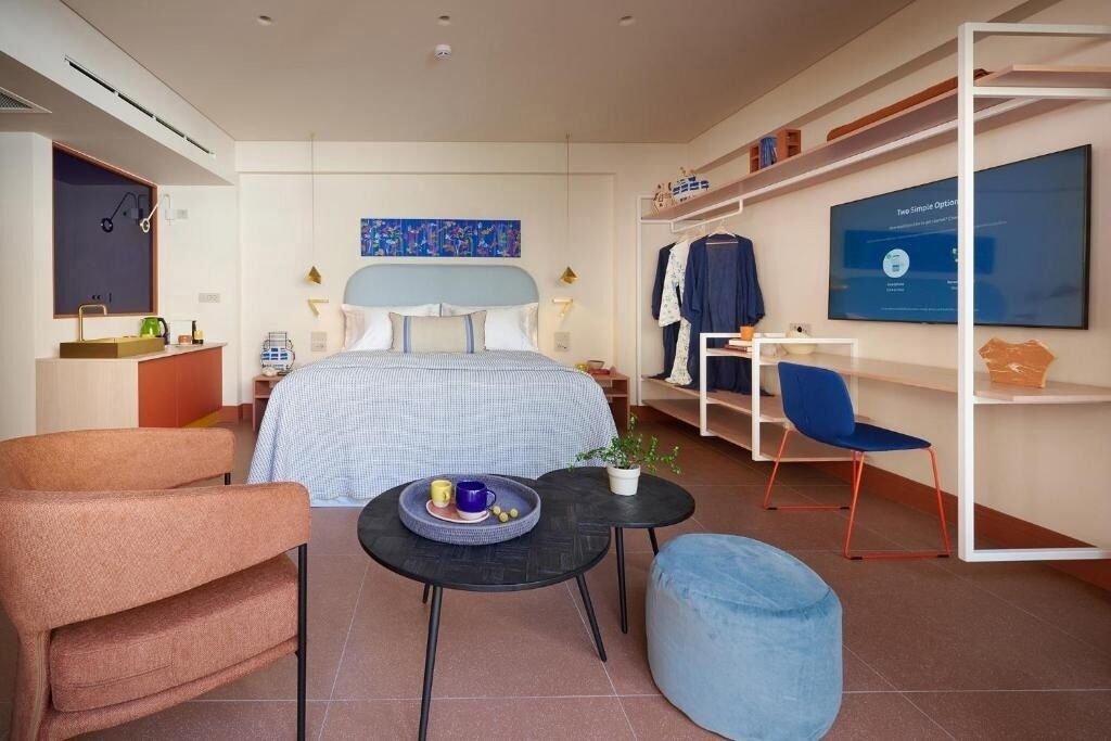Moderni hotellihuone, jossa on sänky, työpöytä, tuoli, pieni tarjoilupöytä ja sininen rahi.