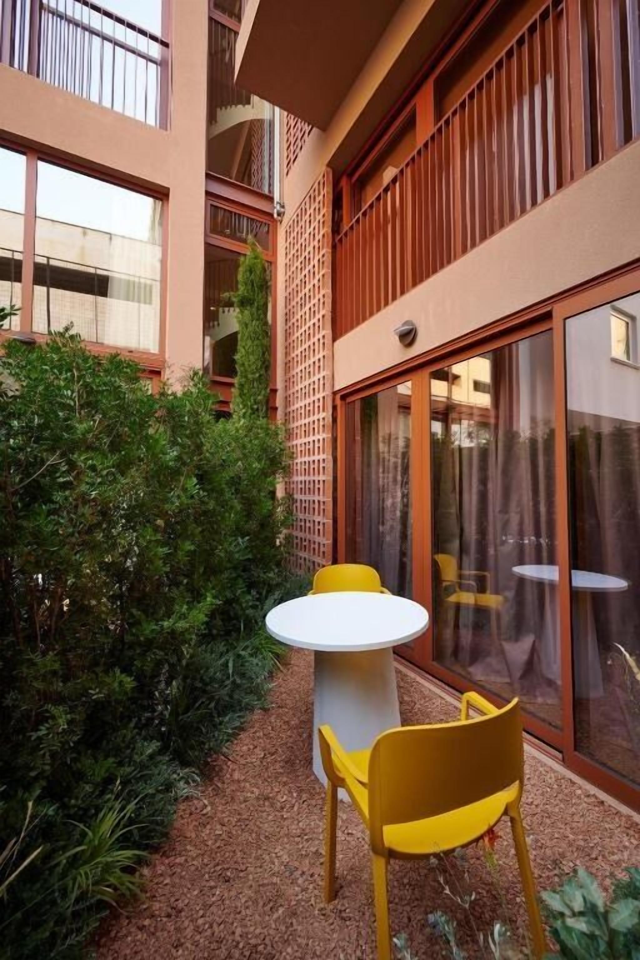 A courtyard with a table and chairs, surrounded by greenery and a building with large windows.