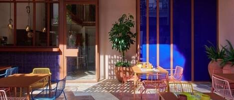 Une terrasse extérieure avec une table et des chaises en bois, un pot de fleurs et une vue sur un bâtiment à travers une porte vitrée.