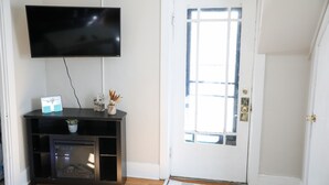 Una habitación con televisión de pantalla plana en la pared, un mueble bajo con una planta y una puerta con ventana.