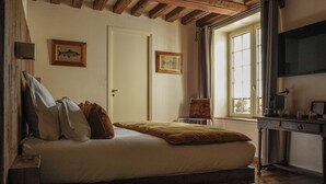 A bedroom with a wooden ceiling, a bed with pillows, a chair, a desk with a lamp, and two framed pictures on the wall. - L'Absinthe Hôtel (Honfleur)