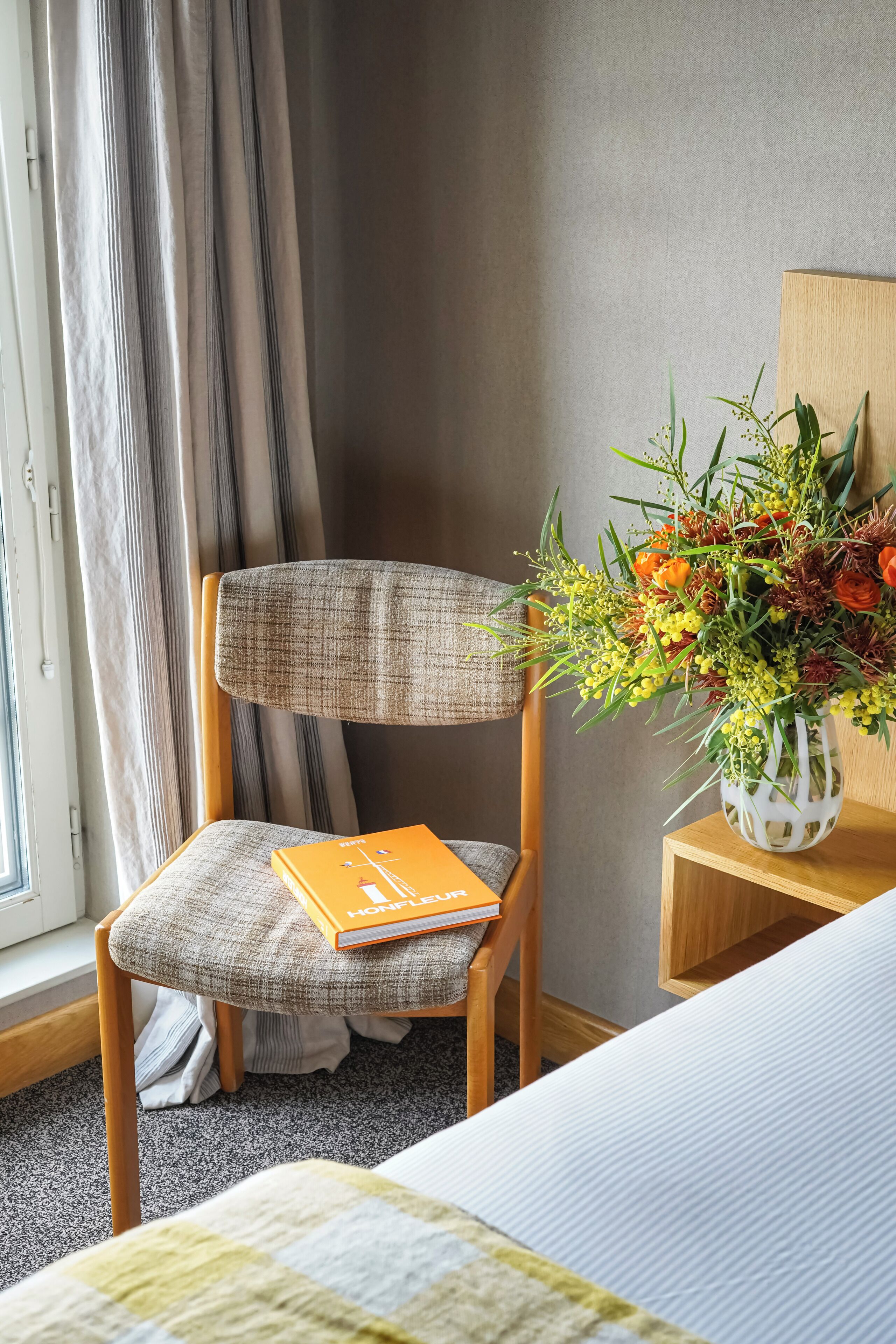 A hotel room with a wooden chair, a bedside table with a vase of flowers, a bed with a quilt, and a window with curtains.