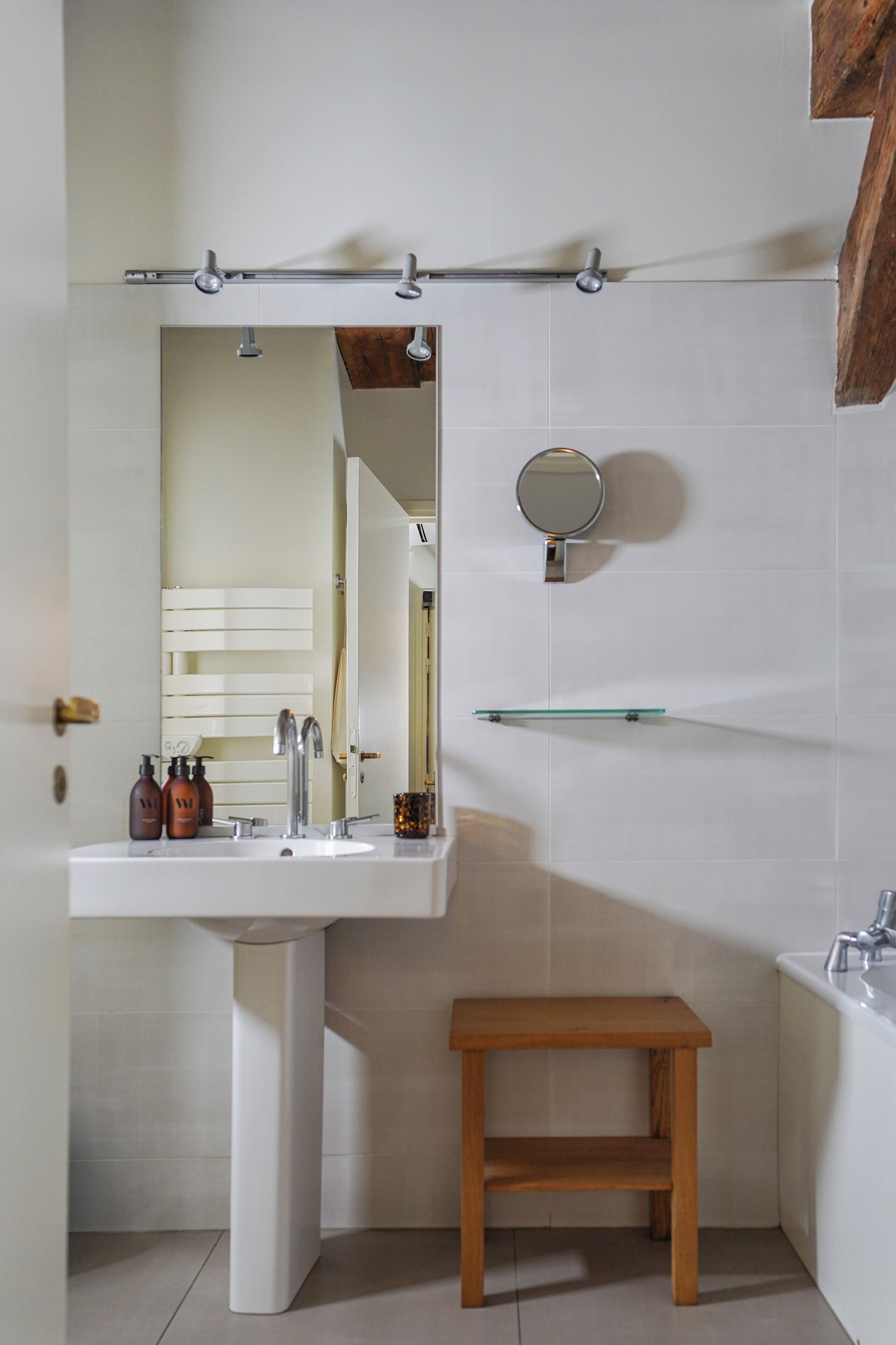 A bathroom with a white pedestal sink, a wooden stool, a mirror, and a glass shelf.