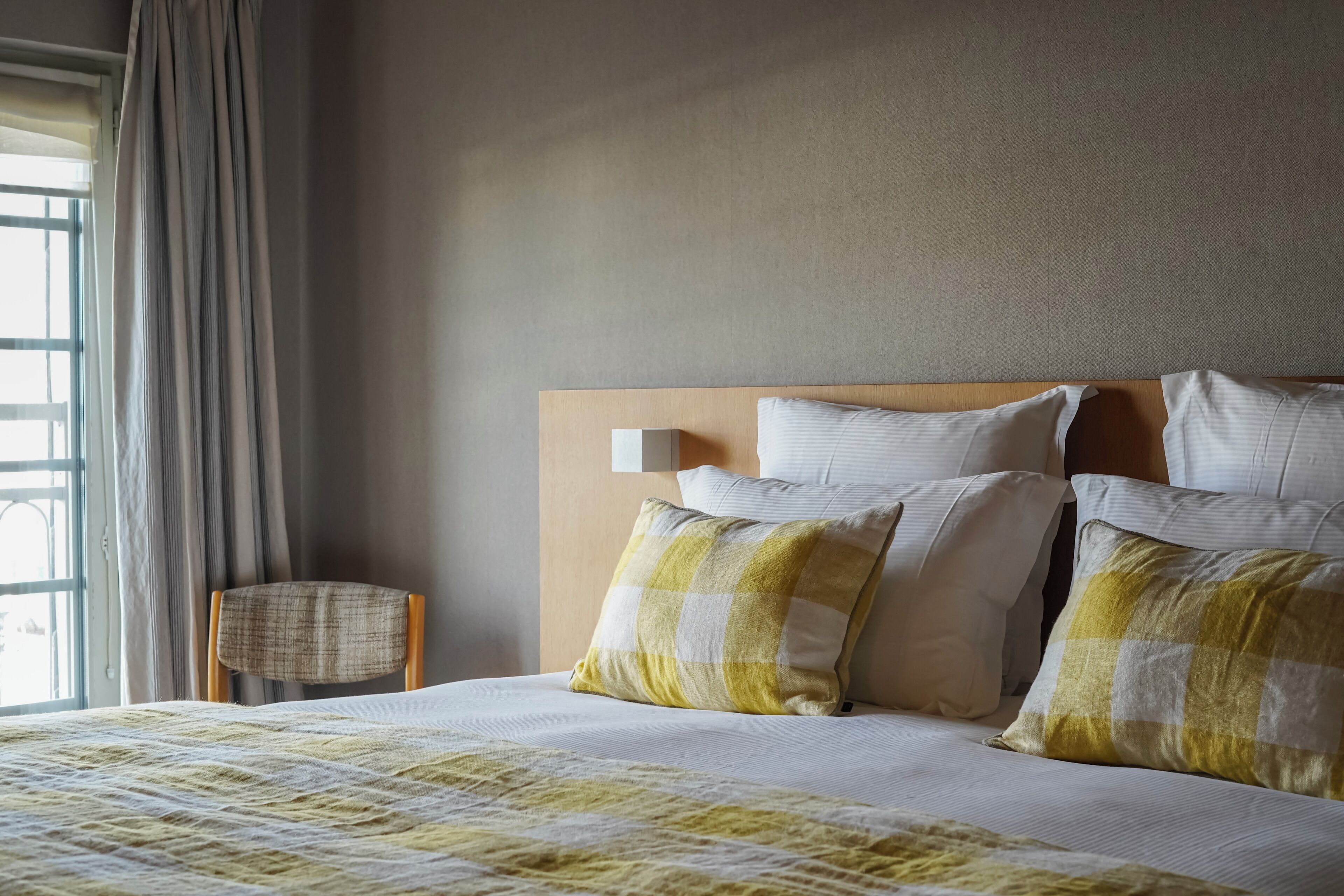A neatly made bed with a checkered pillow, a wooden headboard, and a chair beside the window.