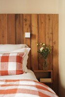 Un dormitorio con cabecera de madera, colcha a cuadros, almohada blanca, un jarrón con flores y una mesita de noche de madera.
