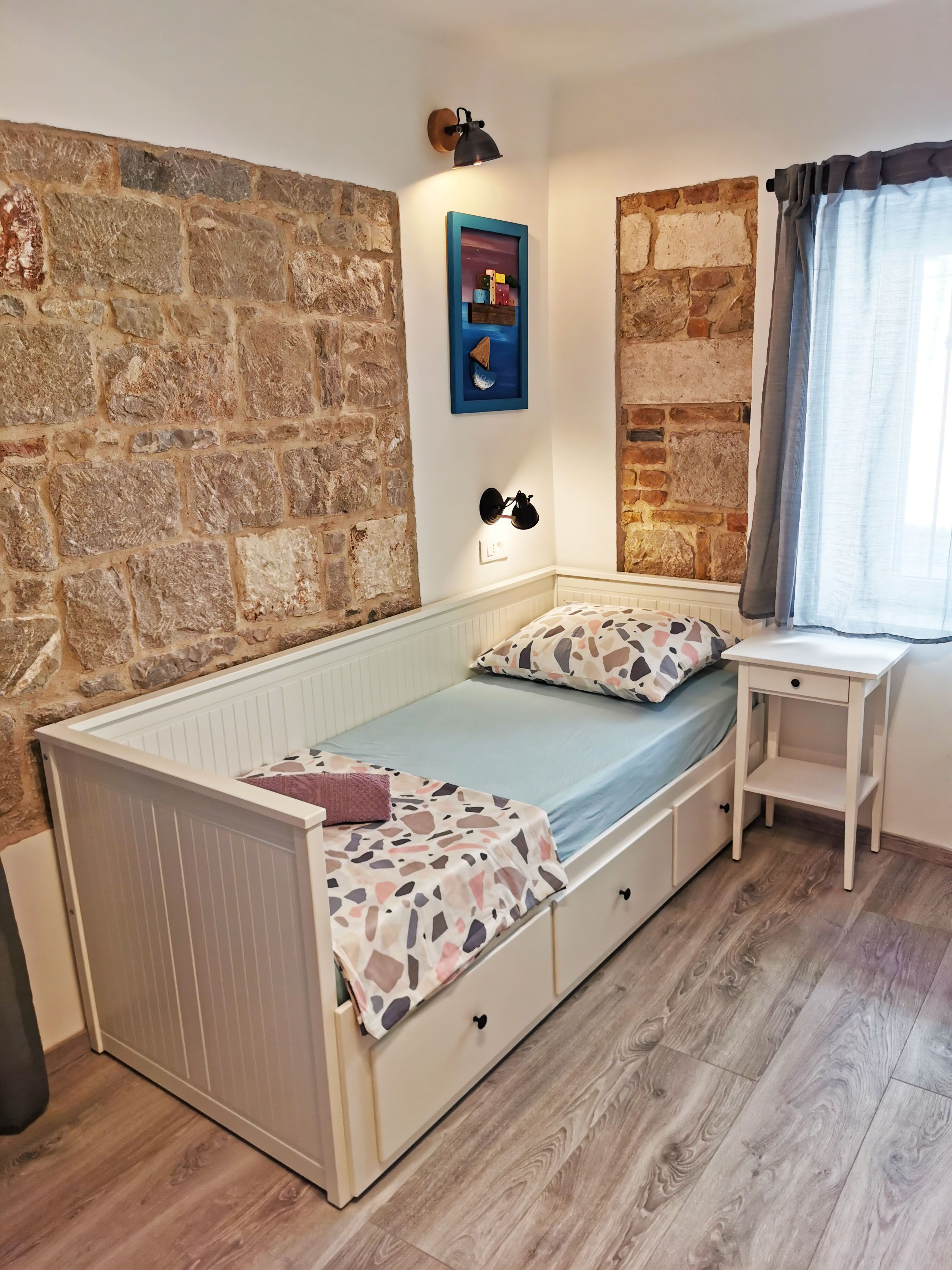 A single-bed room with a stone wall, a small bedside table, a nightstand, and a window with curtains.