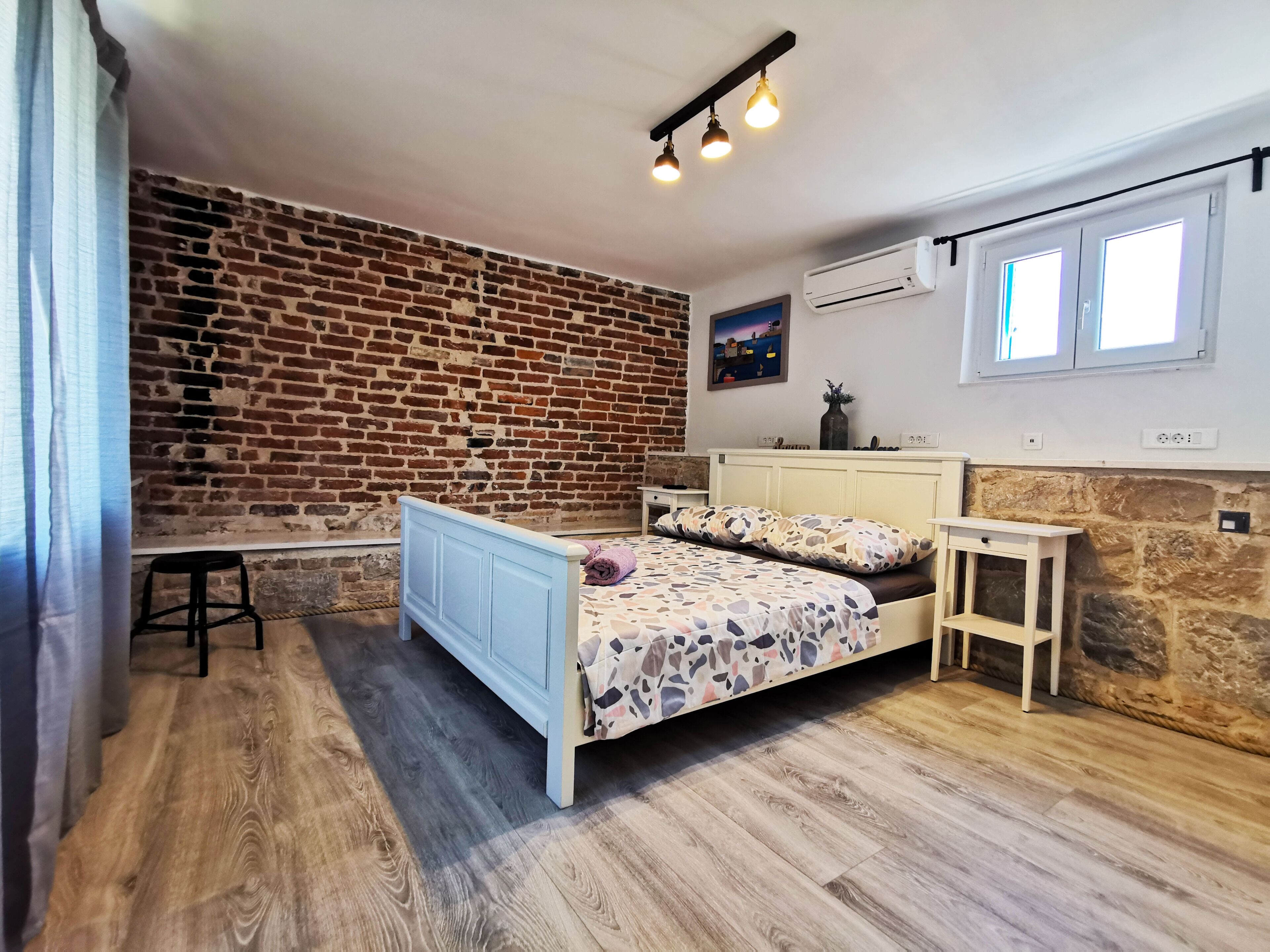 A bedroom with a brick wall, a bed with patterned bedding, a small table, and a window with daylight.