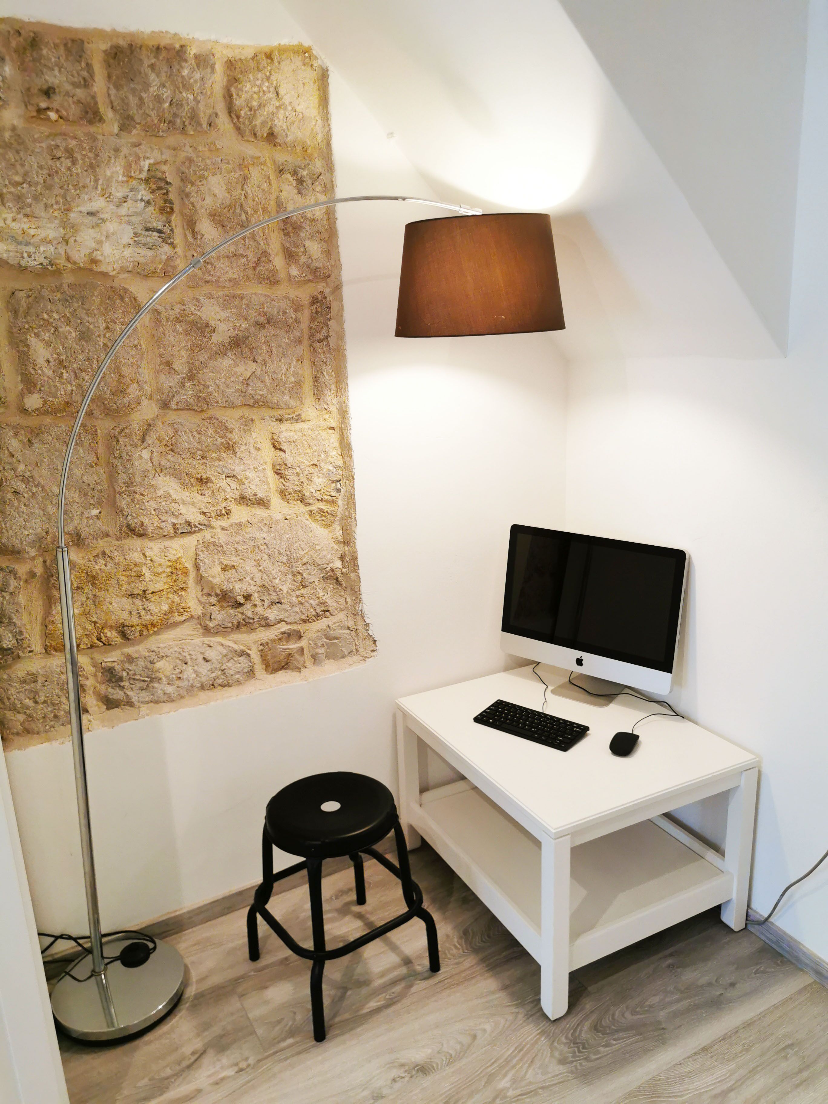 A small, white desk with a computer monitor, keyboard, and mouse. There is a black stool and a floor lamp with a brown shade. The wall is partially covered with a stone-like texture.