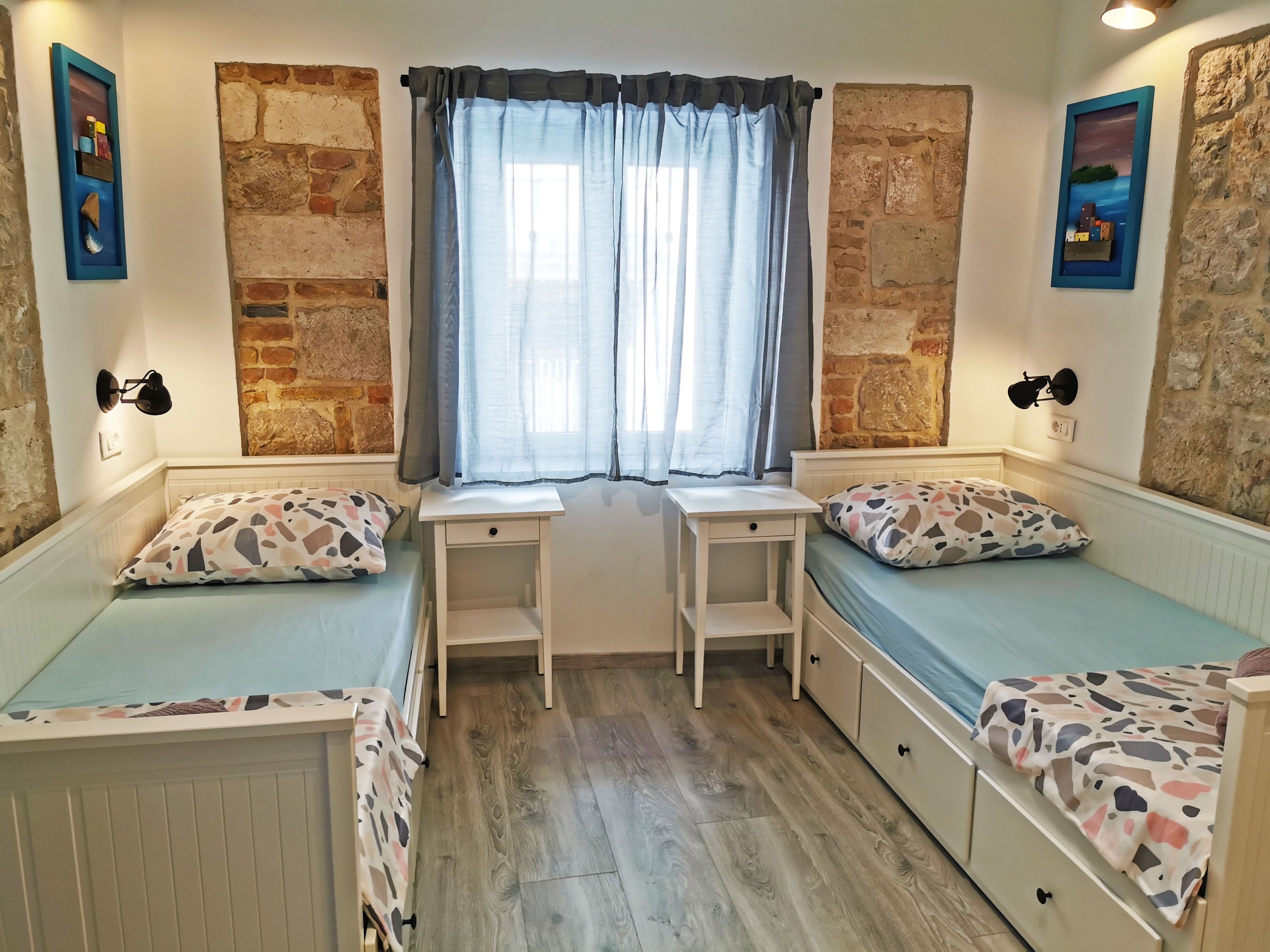 A cozy room with a bed, two small desks, a window with curtains, and stone walls.