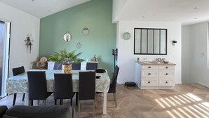 A spacious dining area with a high ceiling and exposed wooden beams, a dining table with chairs, and a kitchen area with a white cabinet and a large mirror.