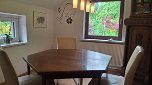 A dining room with a wooden table, chairs, a radiator, a window with a view of greenery, and wall art. - Detached thatched house (78sqm) with a large garden in the middle of the Butjadingen peninsula (Butjadingen)