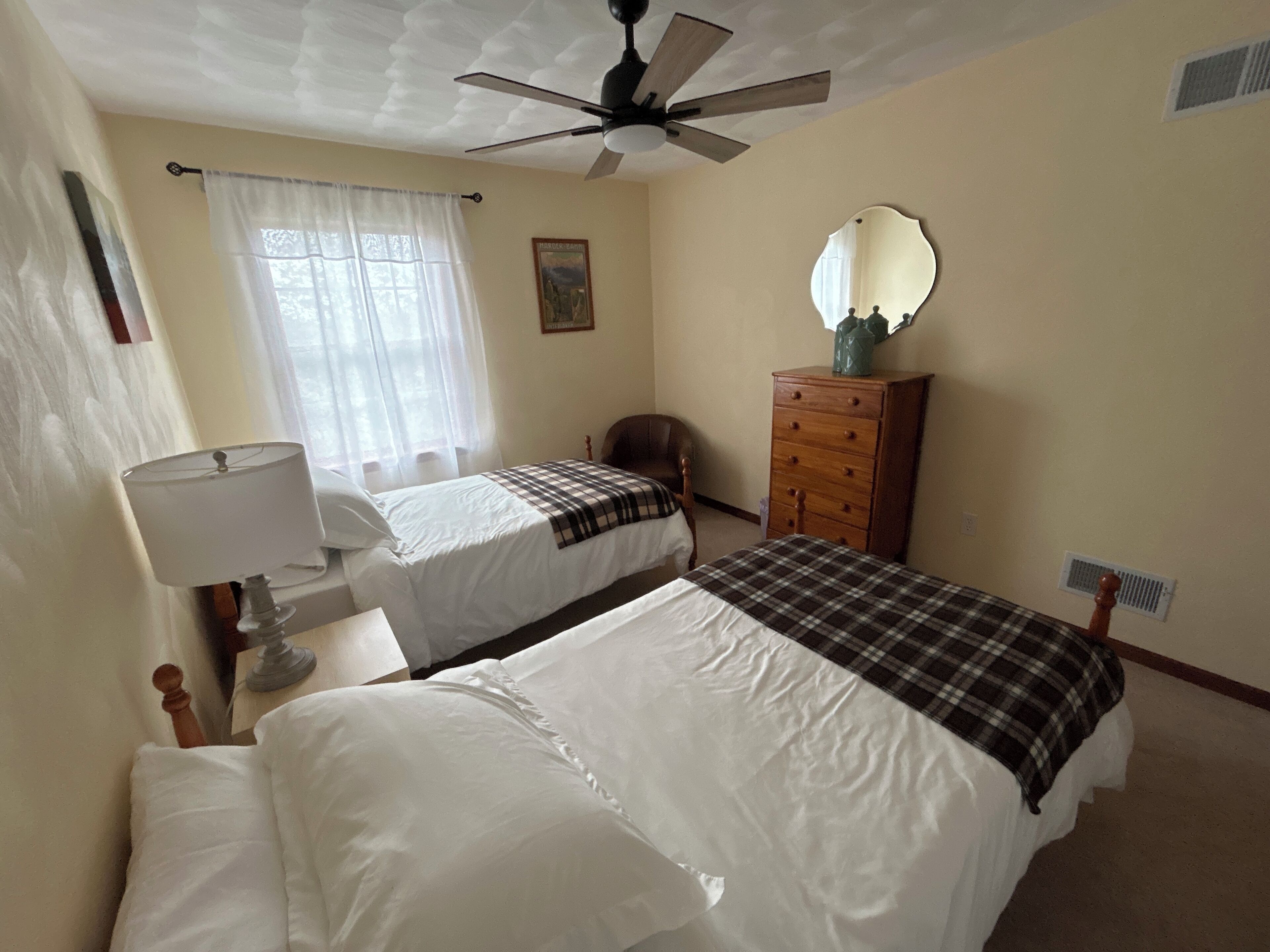 A bedroom with two beds, a ceiling fan, a dresser, a mirror, and a window with curtains.