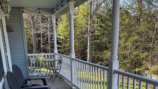 A covered porch with outdoor seating, white railings, and a view of a forested area.