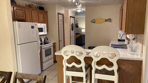 A compact kitchen with wooden cabinets, a white refrigerator, a microwave, and an oven. There are two white chairs with patterned cushions at a small dining table.