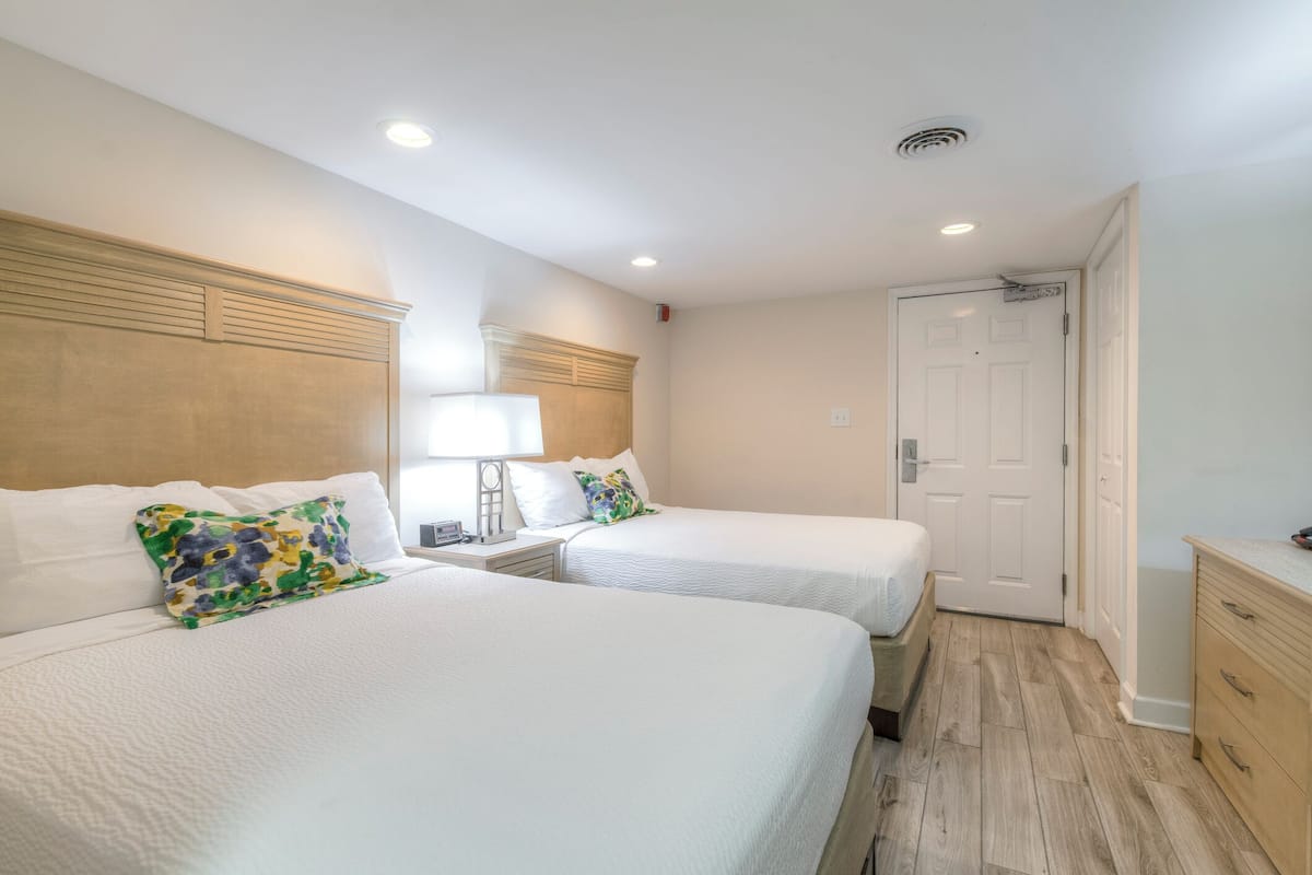 A hotel room with two single beds, a wooden headboard, a bedside table with a lamp, a white door, and a wooden dresser.