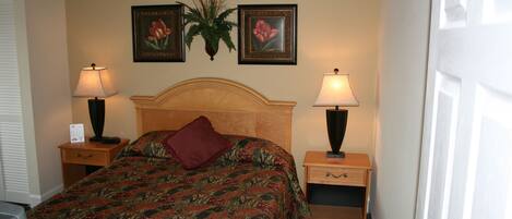 A hotel room with a bed, two bedside tables with lamps, a nightstand, a wall-mounted plant, and two framed pictures.