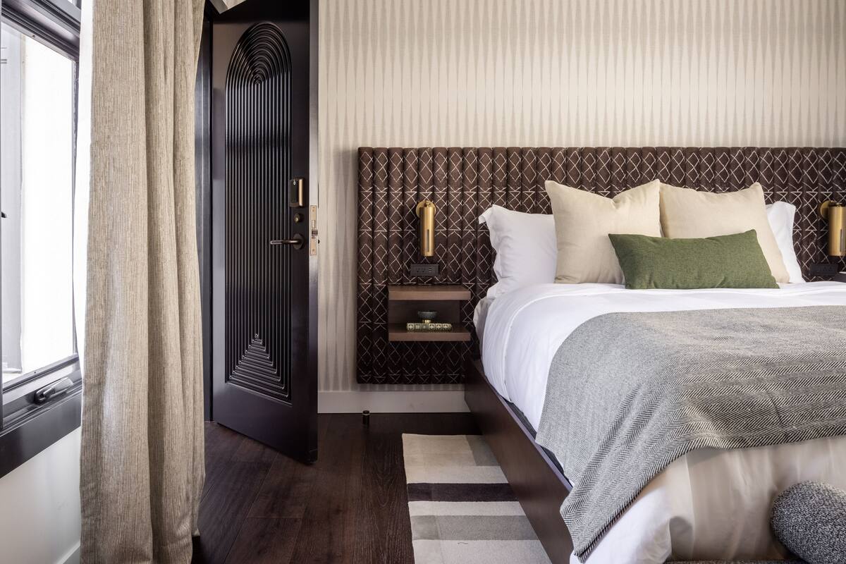 A modern hotel room with a large bed, a dark wood headboard, a nightstand with a lamp, and a view of the outside through a window.