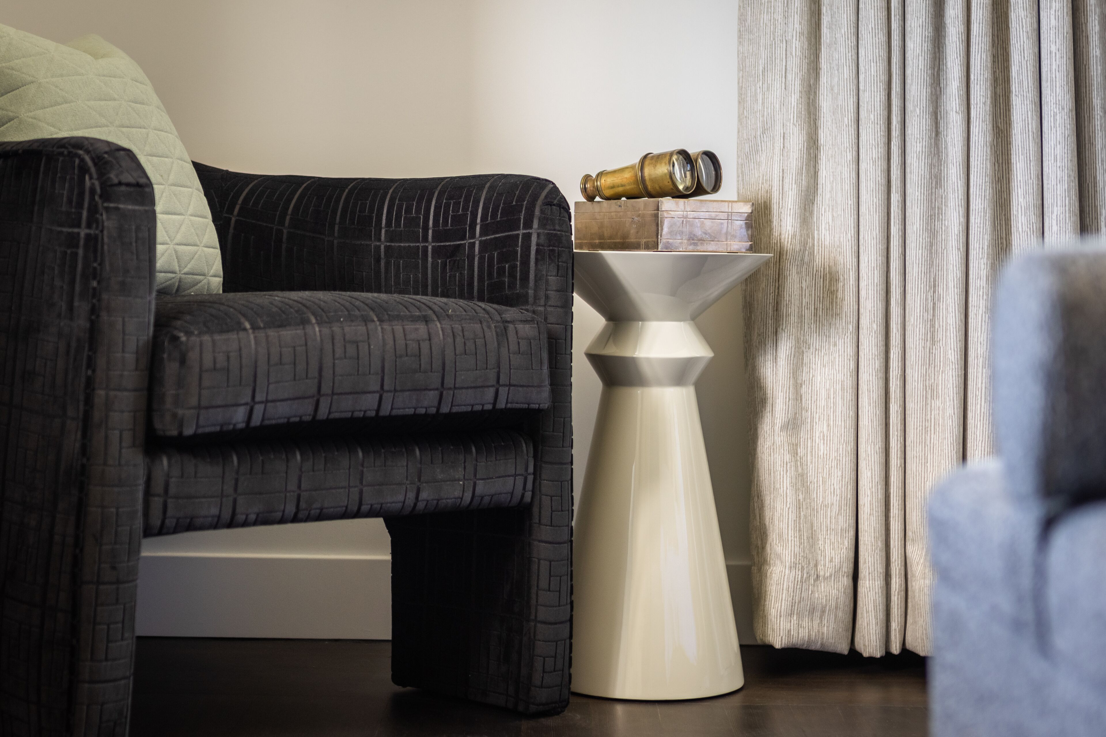 Una sala de estar moderna con un sofá de tono oscuro y estampado, una mesita auxiliar blanca con un objeto decorativo y una cortina.