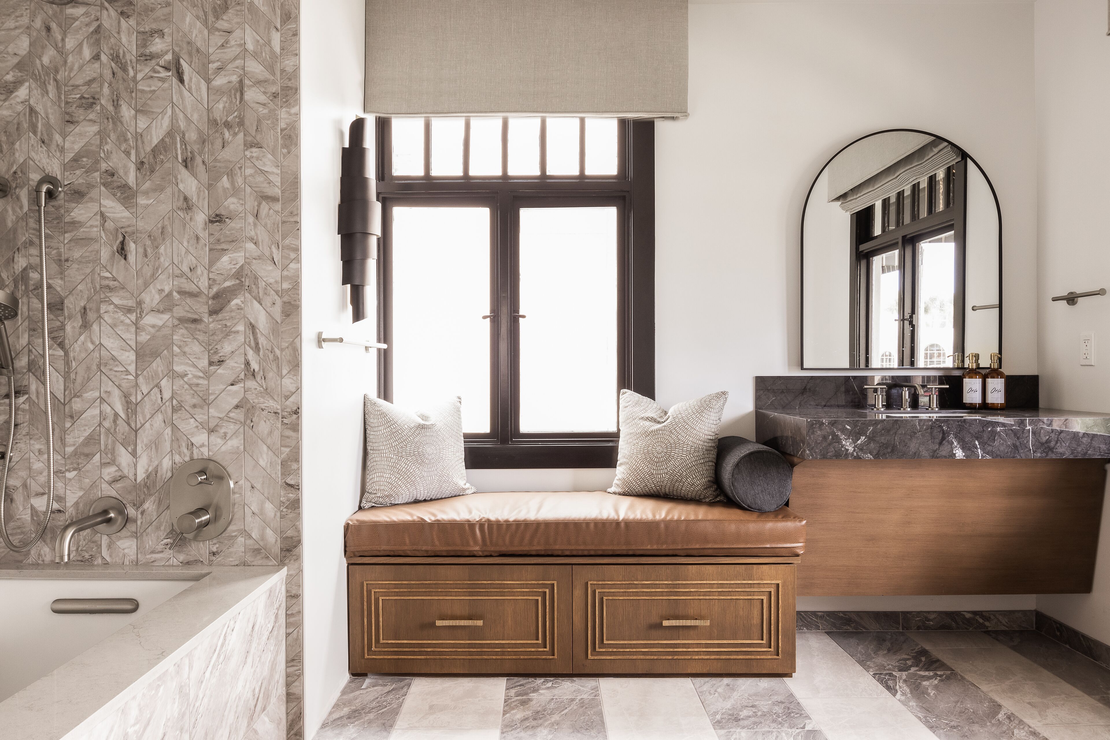 A bathroom with a herringbone tile backsplash, a bathtub, a bench with storage drawers, a vanity with a mirror, and a window with a view.