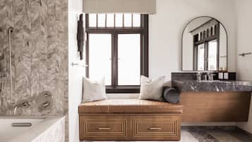 A bathroom with a herringbone tile backsplash, a bathtub, a bench with storage drawers, a vanity with a mirror, and a window with a view.