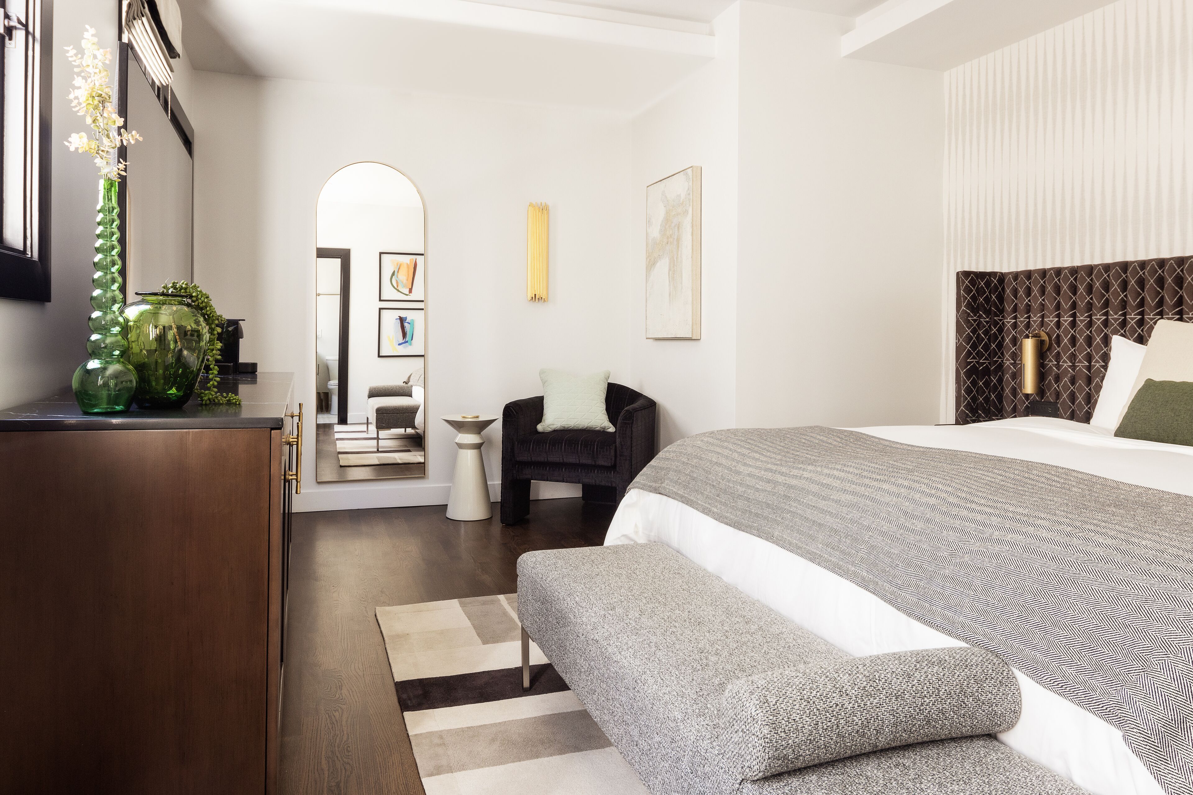 A modern bedroom with a large bed, a bench, a small side table, and a green vase on a dresser.