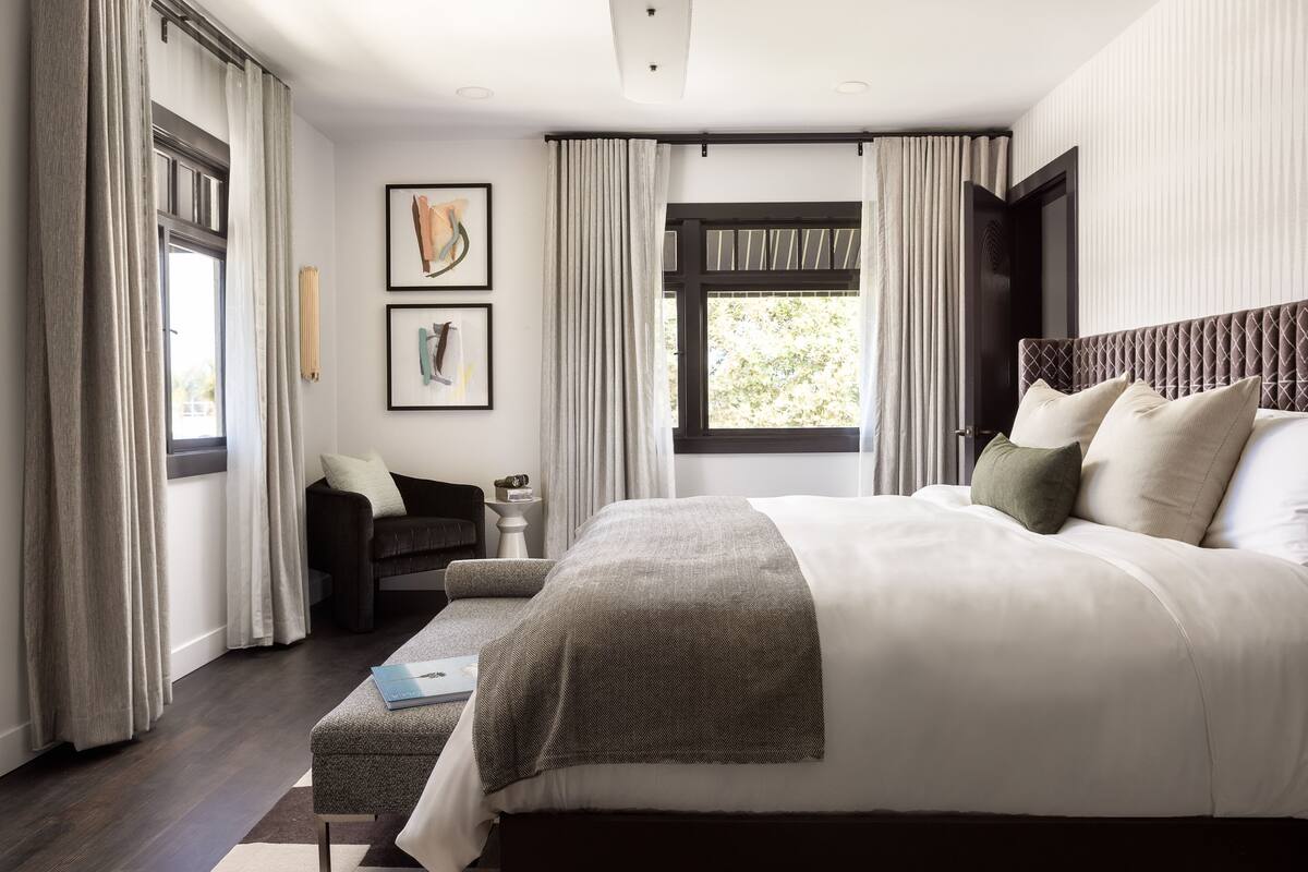 A modern bedroom with a large bed, a bedside table, a reading chair, and a window with curtains.