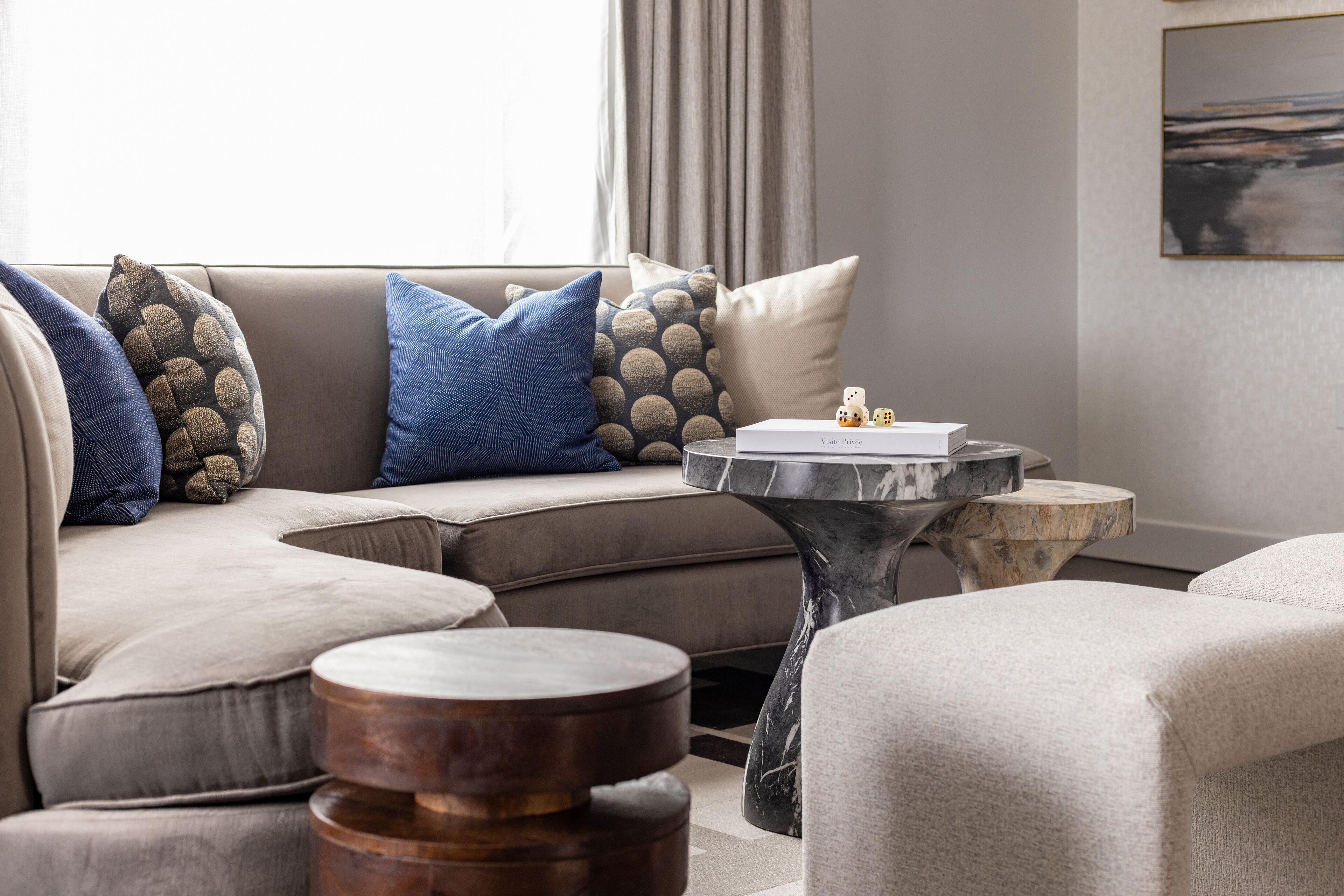 A modern living room with a beige sectional sofa, blue and patterned throw pillows, a marble coffee table, and a small wooden side table.