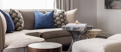 A modern living room with a beige sectional sofa, blue and patterned throw pillows, a marble coffee table, and a small wooden side table.