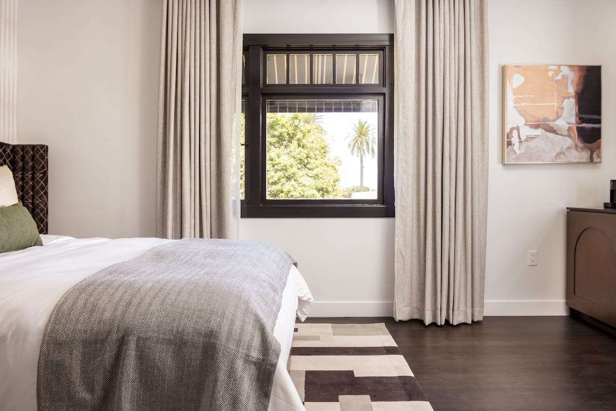 A bedroom with a large bed, a window with curtains, a painting on the wall, and a wooden cabinet.