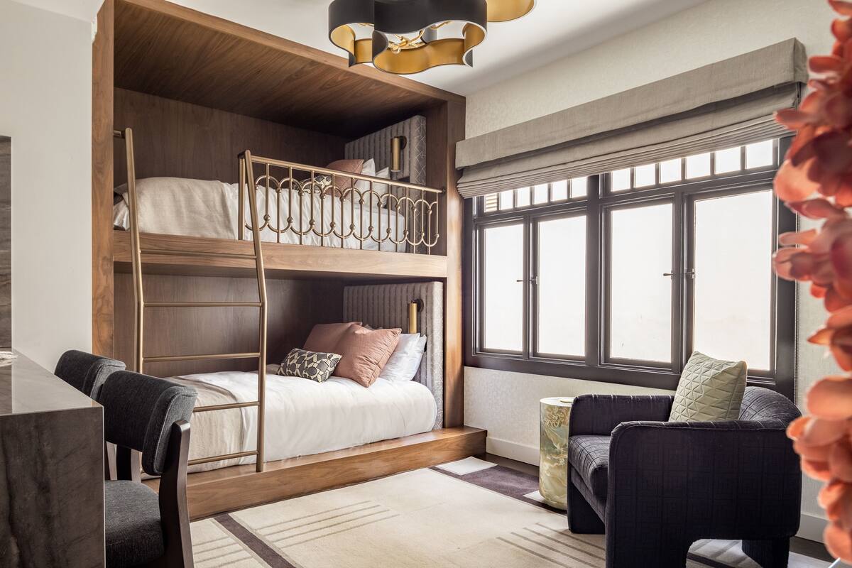 A room with a bunk bed, a desk, a chair, a sofa, and a window with blinds.
