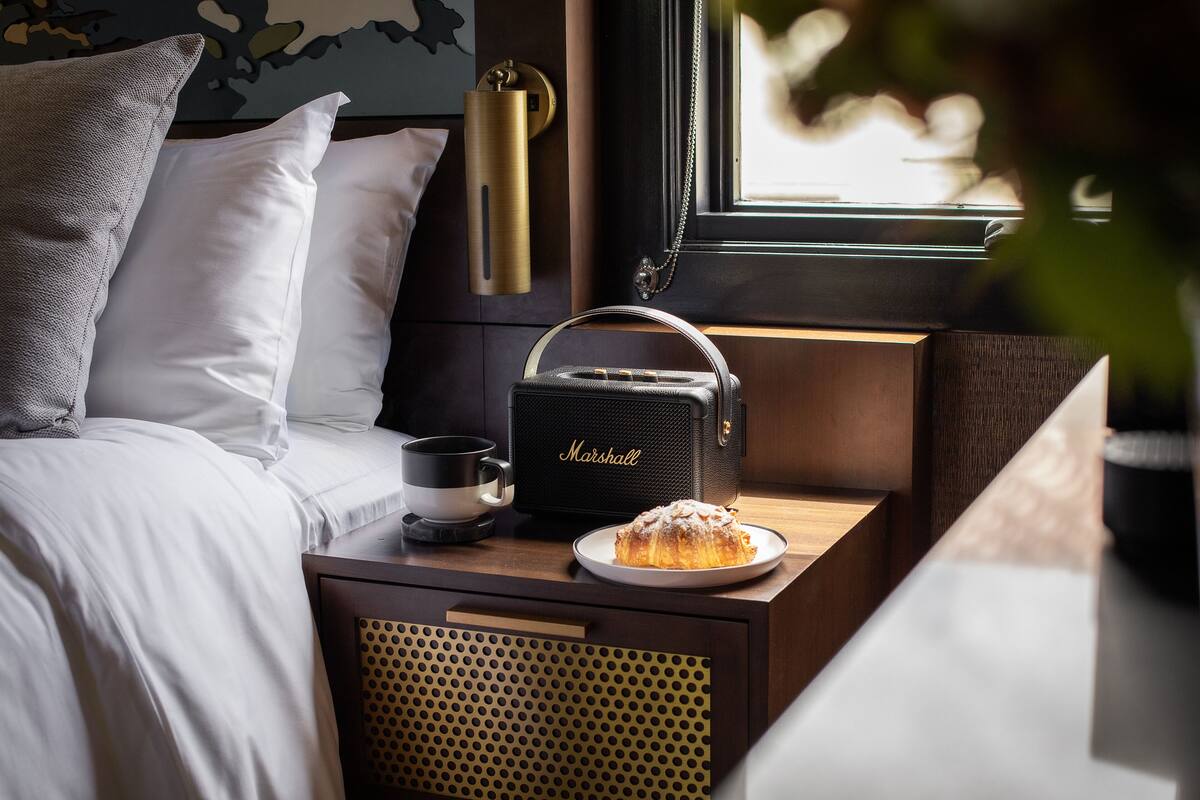 A neatly made bed with white linens, a Marshall portable speaker, a plate with a dessert, and a coffee cup on a bedside table.
