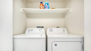 Une buanderie équipée de deux machines à laver et d’un sèche-linge, ainsi que d’une étagère au-dessus où sont posées deux bouteilles de lessive.