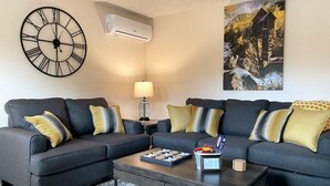 A modern living room with two sofas, a coffee table, a wall clock, an air conditioner, and a painting of a rustic house by a river. - ★Academy Hideaway With View! Sleeps 4 with AC!★ (Colorado Springs)