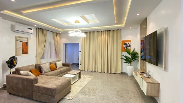 A modern living room with a sectional sofa, a flat-screen TV, a ceiling light fixture, and a potted plant. - TAOhomes (Abuja)