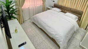 A hotel room with a bed, curtains, a TV, a bedside table, and a plant. - TAOhomes (Abuja)