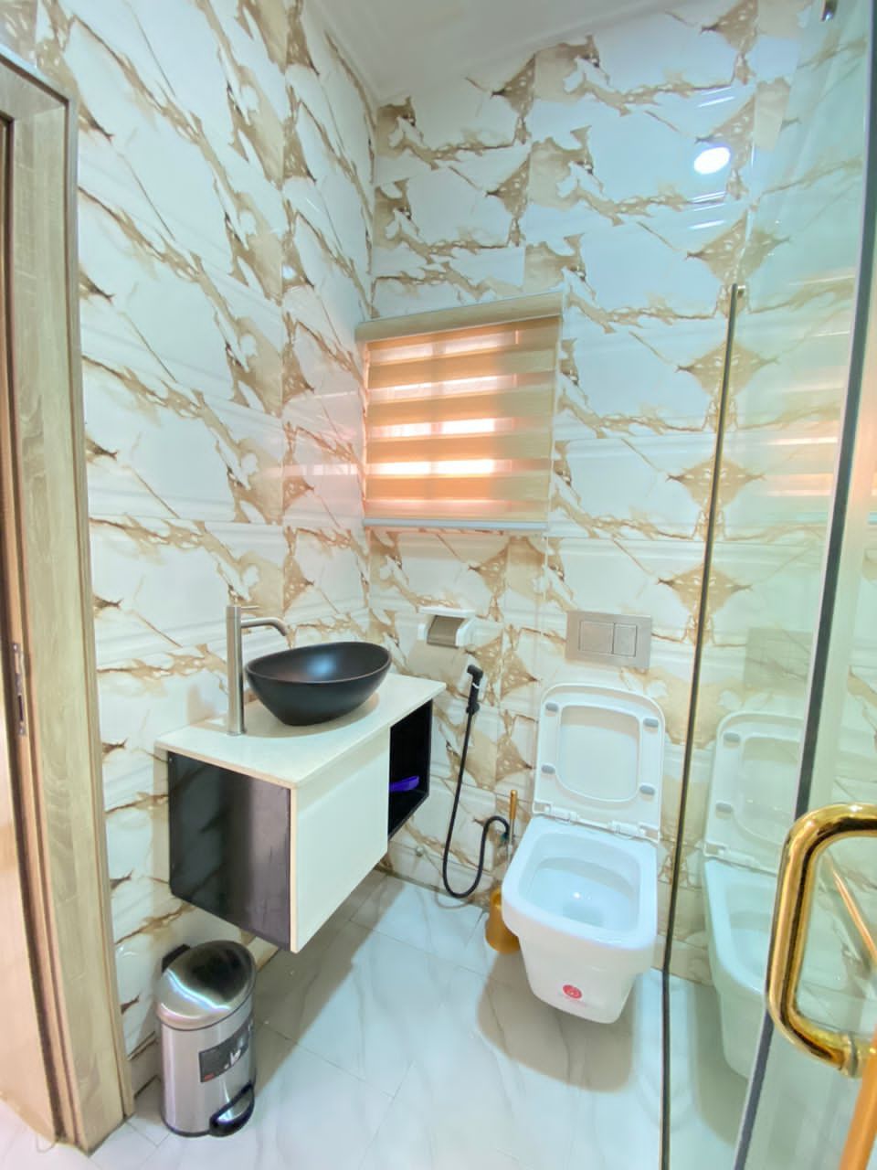 Casa de banho compacta com painel de fundo de mármore, lavatório preto, sanita branca e um contentor de lixo.