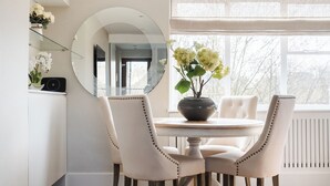 A dining area with a round table, four upholstered chairs, a vase with flowers, a round mirror, and a window with a view of trees. -  LONDONERS│Magnificent Londoner  (London)