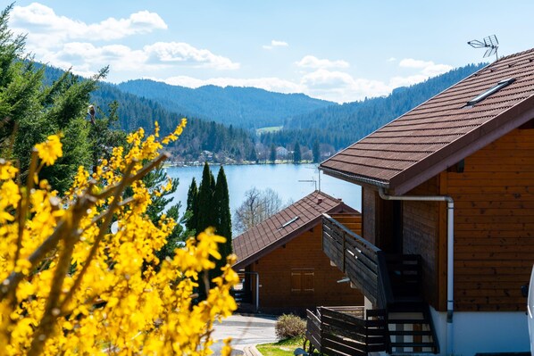 A wooden cabin with a balcony overlooking a lake and forested mountains.