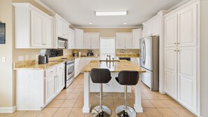 A modern kitchen with white cabinetry, a central island with bar stools, stainless steel appliances, and a large window. - Large spacious Home (Enterprise)